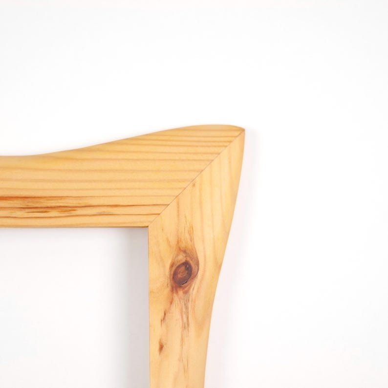 May include: A light-colored wooden frame corner featuring a visible natural wood grain pattern and a distinct knot. The frame has a smooth, polished finish with a mitered joint connecting the horizontal and vertical pieces of light pine wood.