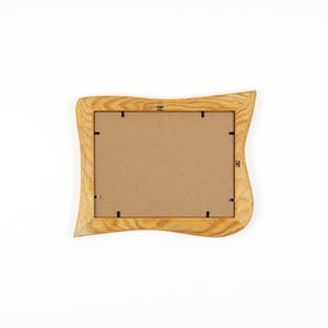 May include: A rectangular picture frame with a light brown wood grain finish and wavy, organic edges. The back of the frame features a flat, tan cardboard backing secured by several small black metal clips and a central metal hanging hook.