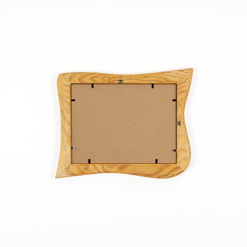 May include: A rectangular picture frame with a light brown wood grain finish and wavy, organic edges. The back of the frame features a flat, tan cardboard backing secured by several small black metal clips and a central metal hanging hook.