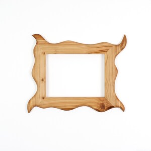 Puede incluir: Un marco de fotos rectangular hecho de madera de color marr&oacute;n claro con un patr&oacute;n de veta natural. El marco presenta un borde exterior irregular y ondulado con esquinas puntiagudas que se extienden hacia afuera, rodeando una abertura central.