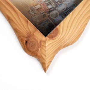 May include: Close-up of a wooden picture frame with a unique, curved design. The frame is made of light-colored wood with visible grain and knots. The frame's interior holds a picture with a blurred interior scene.