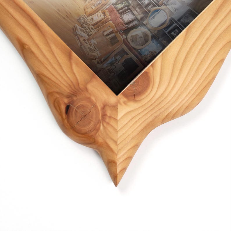 May include: Close-up of a wooden picture frame with a unique, curved design. The frame is made of light-colored wood with visible grain and knots. The frame's interior holds a picture with a blurred interior scene.