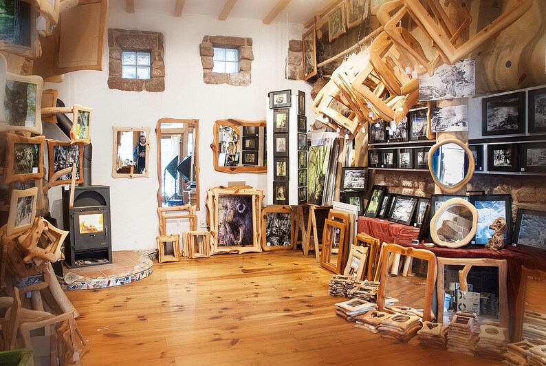 May include: A gallery showcasing framed artwork and mirrors with natural wood frames. The space has a warm atmosphere with wooden floors and a variety of framed pieces on display, including landscape and animal prints. The walls have stone windows.