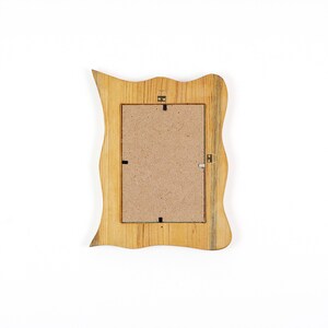 May include: Wooden picture frame with a wavy, irregular edge. The frame is made of light-colored wood with a natural grain pattern. The back of the frame is visible, showing a cardboard backing and hanging hardware.