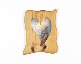May include: A light brown wooden wall mirror with a natural wood grain pattern and visible knots. The frame features an irregular, wavy edge and a central heart-shaped cutout that reveals the reflective glass surface within the wooden structure.