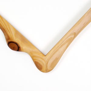 Op de afbeelding: Close-up van een houten kledinghanger. De hanger is gemaakt van lichtgekleurd hout met zichtbare nerf en een knoopdetail. Het ontwerp is een eenvoudige V-vorm, ideaal voor het presenteren van kleding.