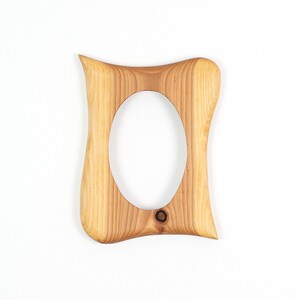 May include: A light-colored wooden picture frame with an oval opening. The frame has a unique, curved shape with rounded edges and a small hole at the bottom. The wood grain is visible.