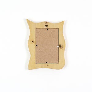 Pu&ograve; includere: Il retro di una cornice portafoto in legno chiaro con un bordo ondulato e sagomato. La cornice presenta un pannello posteriore rettangolare in fibra di legno compressa, fissato da piccole clip metalliche, e include un gancio metallico in alto al centro.