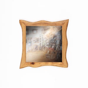 May include: A square wooden picture frame with a wavy edge. The frame is made of light-coloured wood and contains a blurred image of a workshop filled with framed artwork. The frame is set against a white background.