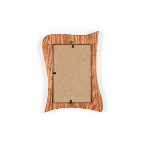 May include: A wooden picture frame with a wavy, decorative edge. The frame is a warm brown color with visible wood grain. The back of the frame has a cardboard backing and a metal hanger for wall display. The frame is empty.