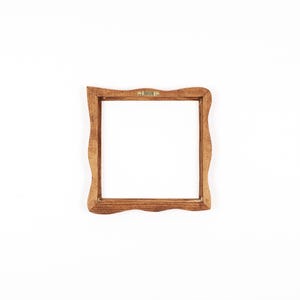 May include: A square wooden picture frame with a medium brown stained finish and wavy, undulating edges on all four sides. The frame features a central open space and a small, gold-coloured metal sawtooth hanger attached to the top inner edge.