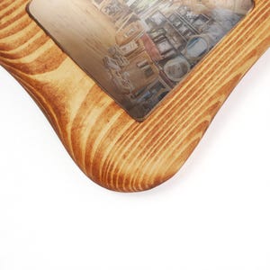 Puede incluir: Un marco de fotos rectangular de madera con esquinas redondeadas, que presenta un patr&oacute;n de veta de madera natural de color marr&oacute;n claro. El marco rodea una fotograf&iacute;a que muestra un espacio interior con varios objetos y muebles.