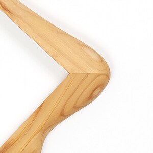 Puede incluir: Una percha de madera de color marr&oacute;n claro con un acabado suave y pulido que muestra patrones de veta de madera natural. La percha tiene una forma curva y ergon&oacute;mica con una uni&oacute;n de hombro angular distintiva y bien definida.