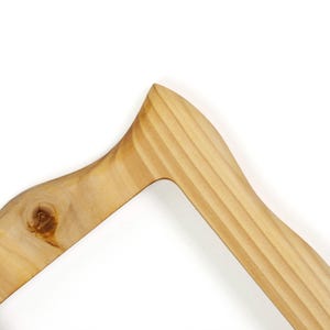 May include: Close-up of a wooden clothes hanger. The hanger is made of light-colored wood with a natural finish. The design features a curved shoulder and a grooved texture on the top portion. A knot is visible on the left side of the hanger.