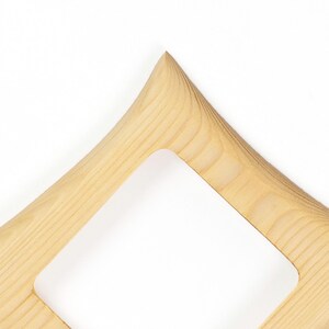 May include: A light-coloured wooden frame with a smooth, natural grain pattern. The frame features a pointed, angular corner design with a square-shaped cutout in the centre, showcasing the light tan wood texture and visible wood grain lines throughout.