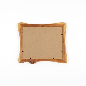 Op de afbeelding: Een houten fotolijst met een natuurlijke houtnerf afwerking. De lijst heeft een unieke, gebogen vorm en een knoopdetail aan de onderkant. De achterkant van de lijst is zichtbaar, met een kartonnen achterkant en metalen ophangmateriaal.
