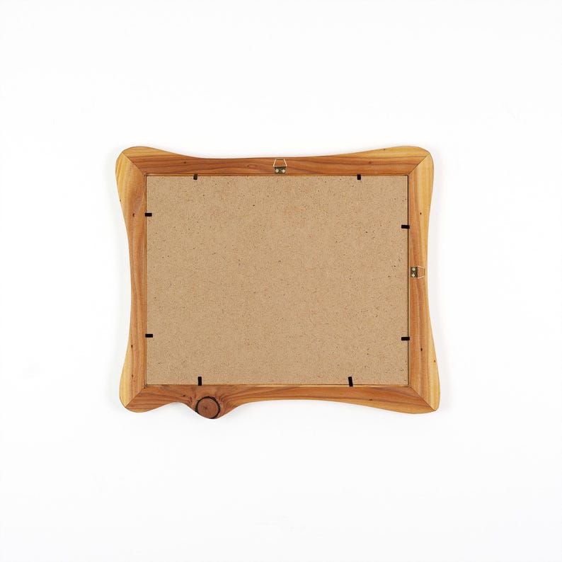 Op de afbeelding: Een houten fotolijst met een natuurlijke houtnerf afwerking. De lijst heeft een unieke, gebogen vorm en een knoopdetail aan de onderkant. De achterkant van de lijst is zichtbaar, met een kartonnen achterkant en metalen ophangmateriaal.
