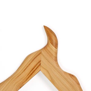 Puede incluir: Una percha de madera con un acabado suave de color marr&oacute;n claro y patrones de veta de madera natural visibles. La percha tiene un dise&ntilde;o de hombro distintivo, curvado y apuntando hacia arriba que se convierte en una estructura de brazo inclinada.