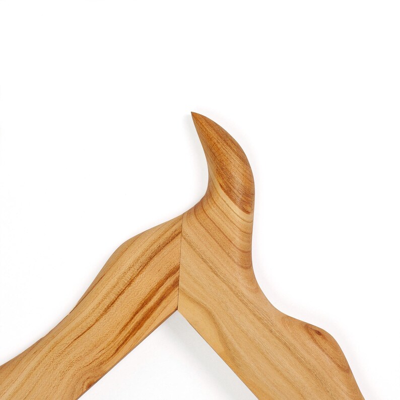 Puede incluir: Una percha de madera con un acabado suave de color marr&oacute;n claro y patrones de veta de madera natural visibles. La percha tiene un dise&ntilde;o de hombro distintivo, curvado y apuntando hacia arriba que se convierte en una estructura de brazo inclinada.