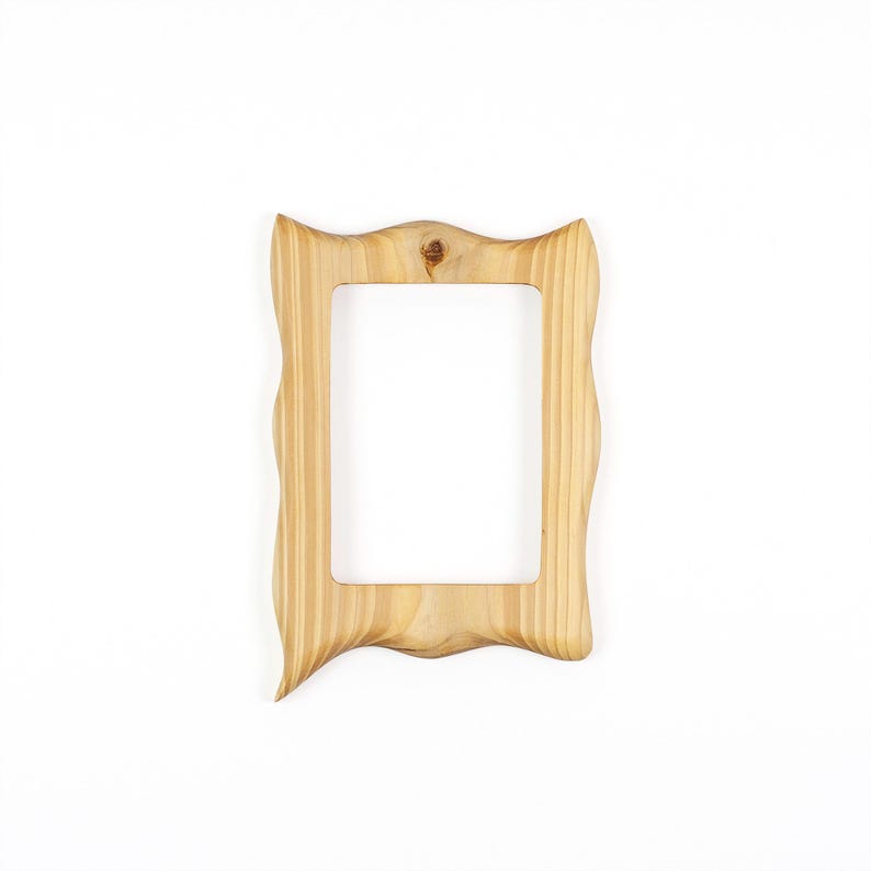 May include: A light-colored wooden picture frame with a wavy, ornate design. The frame has a rectangular opening and a natural wood grain finish. The frame is set against a white background.