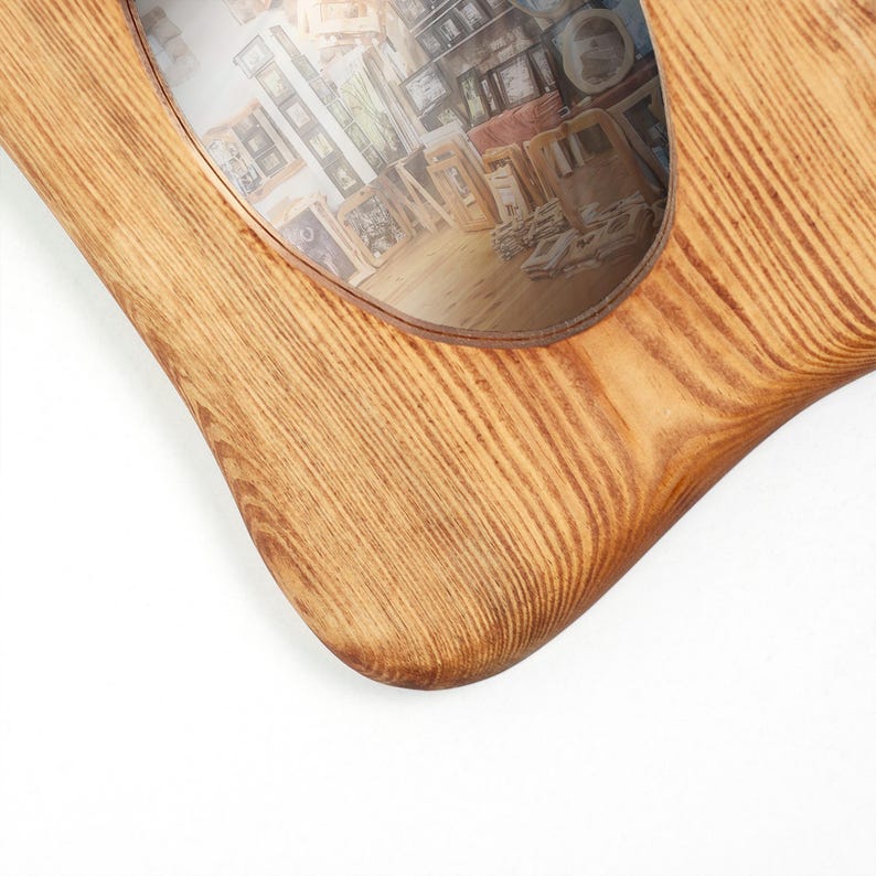 May include: A light brown wooden picture frame with a prominent natural wood grain pattern and a smooth, rounded corner. The frame features an oval-shaped opening that displays a photograph of various other picture frames arranged in a shop.