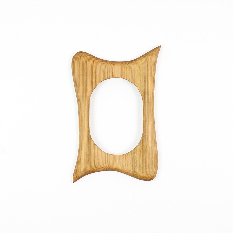 Peut inclure: Un cadre photo en bois brun clair avec une finition lisse et des motifs de grain naturel visibles. Le cadre pr&eacute;sente une silhouette irr&eacute;guli&egrave;re et incurv&eacute;e avec une ouverture ovale centrale et un coin sup&eacute;rieur pointu, r&eacute;v&eacute;lant une texture bois&eacute;e.