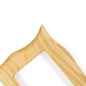 May include: A light brown wooden frame with a smooth, polished finish and a distinct grain pattern. The frame features a sharp, pointed corner at the top and curved, flowing edges that create an open, angular shape with a hollow center.