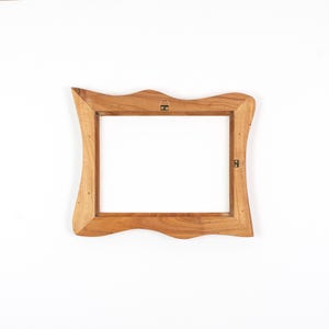 Op de afbeelding: Een houten fotolijst met een uniek, golvend randontwerp. De lijst is gemaakt van lichtbruin hout en heeft een rechthoekige opening. Het heeft een metalen hanger aan de achterkant voor wandmontage.