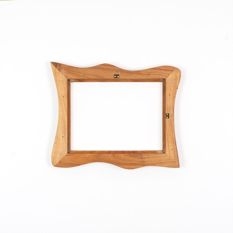 Op de afbeelding: Een houten fotolijst met een uniek, golvend randontwerp. De lijst is gemaakt van lichtbruin hout en heeft een rechthoekige opening. Het heeft een metalen hanger aan de achterkant voor wandmontage.