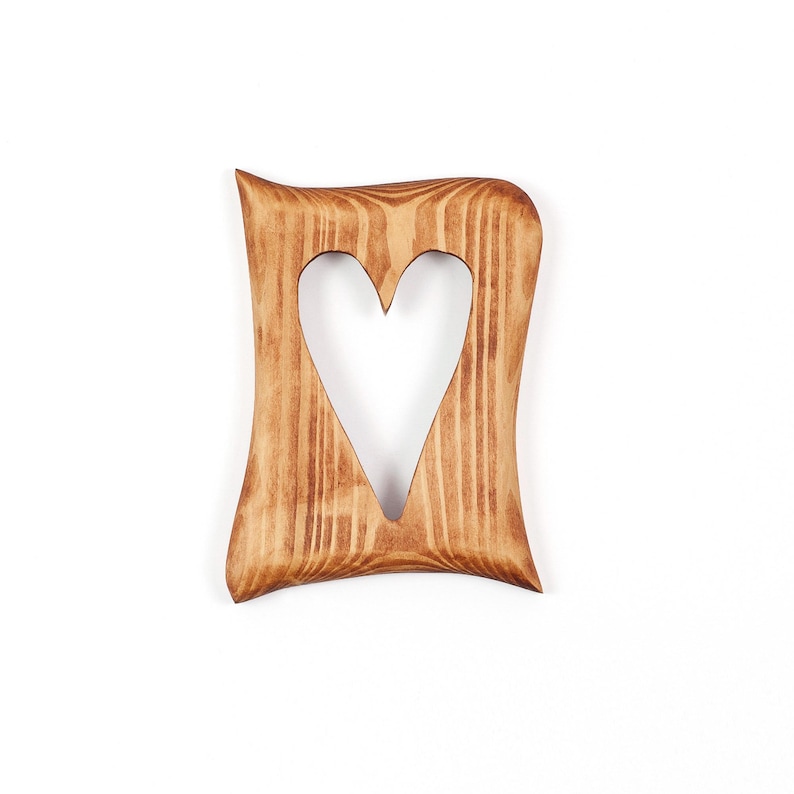 May include: A wooden frame with a heart-shaped cutout. The frame is made of light brown wood with a natural grain pattern. The edges of the frame have a wavy design, and the heart is centered.