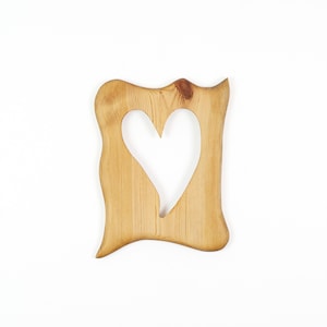 Op de afbeelding: Een lichtbruin houten frame met een natuurlijk houtnerfpatroon en een gladde afwerking. Het frame heeft een onregelmatige, golvende buitenrand en een centrale uitsparing in de vorm van een hart, wat de organische textuur van het hout toont.