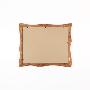 May include: A rectangular picture frame with a natural, wavy wooden edge and a light brown wood grain finish. The back of the frame is covered by a flat, tan-coloured backing board secured with small metal clips and includes two metal hanging hooks.