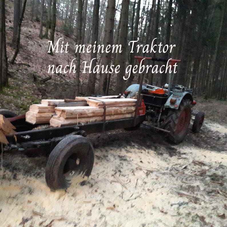 Peut inclure: Un tracteur bleu tirant une remorque noire charg&eacute;e de planches de bois clair empil&eacute;es dans une for&ecirc;t. Le texte "Mit meinem Traktor nach Hause gebracht" est superpos&eacute; en &eacute;criture blanche sur la partie sup&eacute;rieure de l'image.