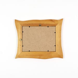 May include: A wooden picture frame with a light brown, wavy edge. The back of the frame is visible, showing a cardboard backing and metal hanging hardware. The frame is set against a white background.