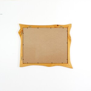 May include: A rectangular wooden picture frame with a natural light brown finish and wavy, organic edges. The back of the frame is covered by a flat, tan-coloured backing board secured with metal clips and includes two metal hanging brackets.