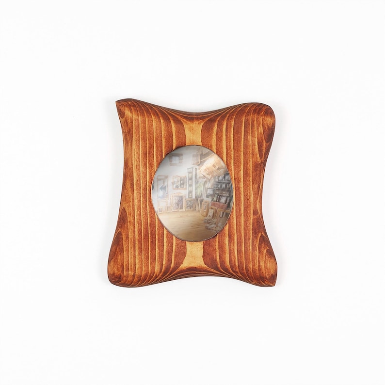 May include: A wooden picture frame with a distinctively curved, four-sided shape and a warm, medium-brown wood grain finish. The frame features a central oval opening that displays a small, blurry photograph of an indoor room with shelves.