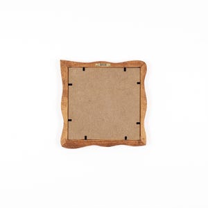 May include: A square wooden picture frame with a wavy, scalloped edge design. The back of the frame features a light brown fibreboard backing secured with eight small black metal clips and a central metal hanging bracket at the top edge.