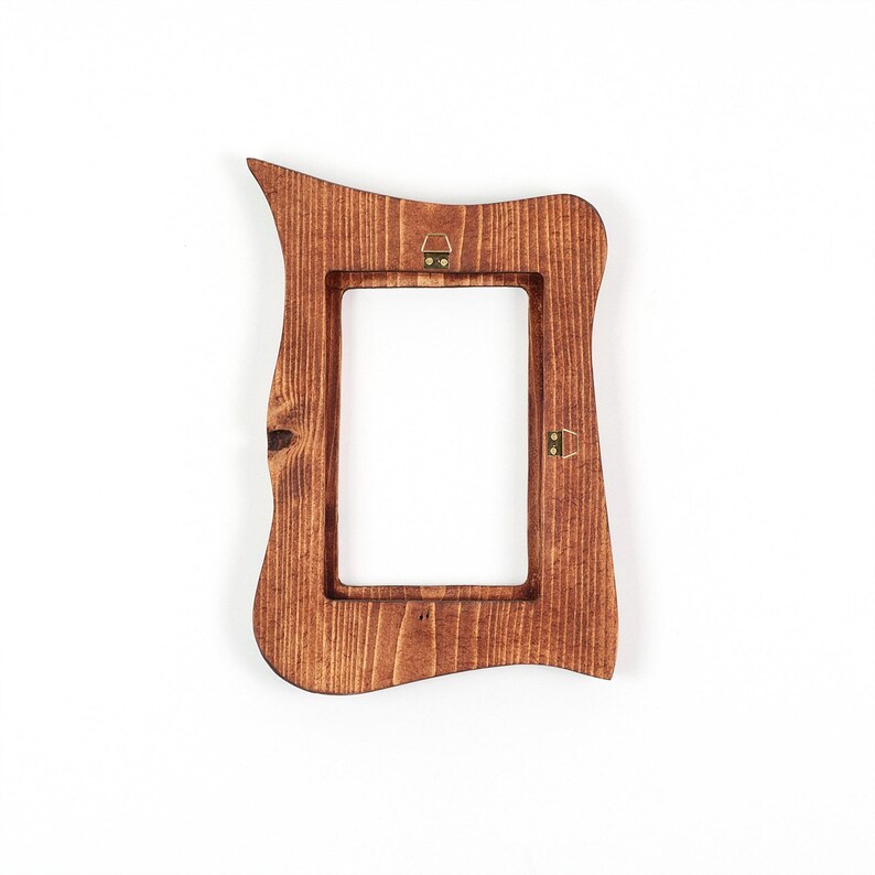 Peut inclure: Un cadre photo en bois teint&eacute; brun moyen avec un bord ext&eacute;rieur irr&eacute;gulier et ondul&eacute; et une ouverture int&eacute;rieure rectangulaire. Le cadre pr&eacute;sente des motifs de grain de bois visibles et deux petites attaches m&eacute;talliques couleur laiton.