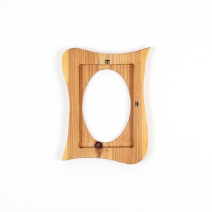May include: A wooden picture frame with an oval opening. The frame is light brown with a wavy, organic shape. It has a natural wood grain texture and metal hanging hardware on the back.