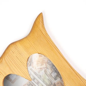 May include: A light-colored wooden mirror with a unique, curved shape. The frame has a natural wood grain pattern and a smooth finish. The mirror's shape is asymmetrical, with a pointed top and a heart-shaped cutout.