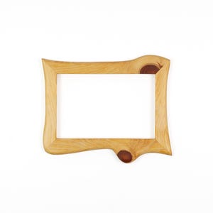 Puede incluir: Un marco de fotos rectangular de madera con un acabado natural marr&oacute;n claro y veta de madera visible. El marco presenta un borde exterior irregular y org&aacute;nico con dos nudos de color marr&oacute;n oscuro integrados en la estructura en la parte superior derecha e inferior.