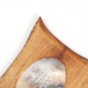 May include: A wooden picture frame featuring a natural light brown wood grain pattern and a distinct pointed top corner. The frame has a smooth, curved cutout opening in the center, showcasing a photograph of various other wooden frames.