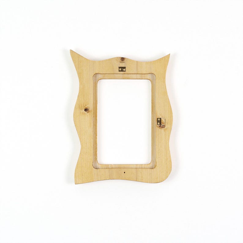 Pu&ograve; includere: Una cornice portafoto in legno chiaro con una silhouette ondulata e organica. La cornice presenta un'apertura centrale rettangolare e include due piccole staffe di montaggio in metallo fissate sul retro per appenderla.