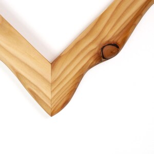 Puede incluir: Una esquina de marco de madera en forma de V con un patr&oacute;n de veta de madera marr&oacute;n claro natural. La pieza muestra un nudo circular oscuro distintivo en el lado derecho y un acabado suave y pulido con una uni&oacute;n a inglete n&iacute;tida en el centro.