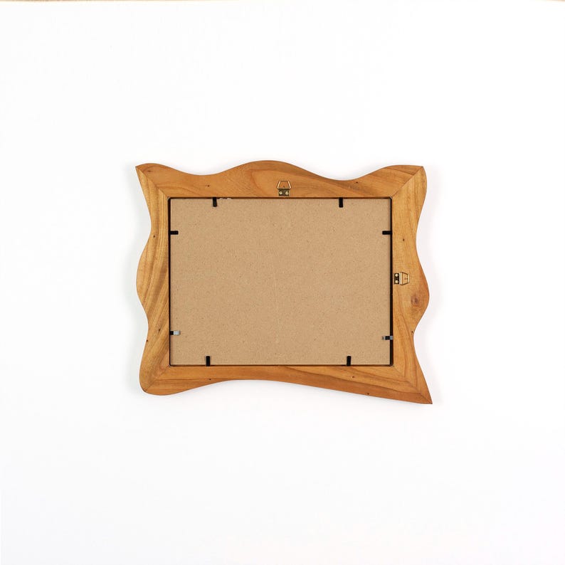 Puede incluir: Un marco de fotos de madera marr&oacute;n claro con un borde decorativo ondulado. El marco tiene una parte trasera de cart&oacute;n y herrajes met&aacute;licos para colgar y sujetar una foto. El marco est&aacute; vac&iacute;o.
