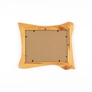 Peut inclure: Un cadre photo en bois brun clair avec un design ondul&eacute; unique. Le cadre pr&eacute;sente un grain de bois naturel et une ouverture rectangulaire pour une photo. Le dos du cadre est visible, montrant le carton et le mat&eacute;riel de suspension.