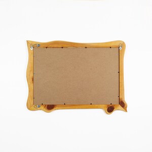 May include: A rectangular wooden picture frame with a natural light brown finish and visible wood grain knots. The back of the frame is covered by a flat brown fiberboard panel secured with metal clips and includes two metal D-ring hangers.