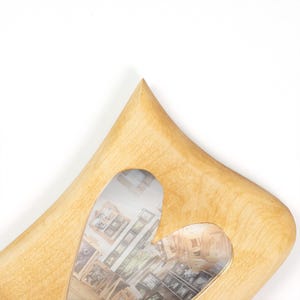 May include: A light-colored wooden picture frame with a heart-shaped cutout in the center. The frame has a smooth, natural grain finish and a pointed top edge. A photograph showing a collection of various framed pictures is visible inside.
