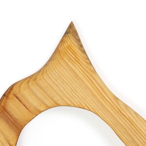 May include: Close-up of a wooden object with a curved, open design. The light-colored wood grain is visible, with a pointed top and rounded edges. The object is set against a white background.
