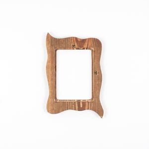 Peut inclure: Un cadre photo rectangulaire en bois avec un bord ext&eacute;rieur ondul&eacute; et irr&eacute;gulier, pr&eacute;sentant un grain de bois naturel visible dans une teinte brun moyen. Le cadre poss&egrave;de une ouverture centrale rectangulaire et des fixations m&eacute;talliques.